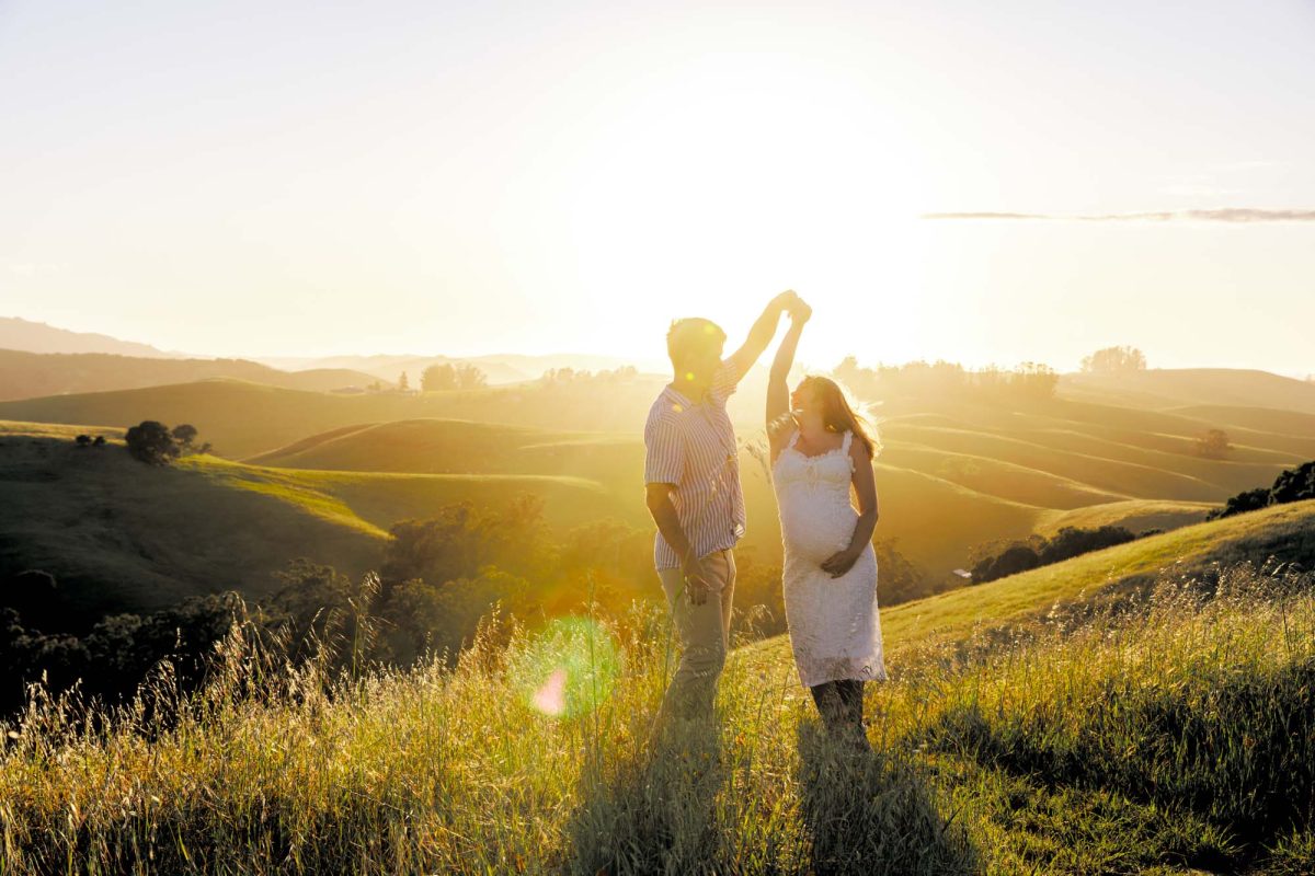 A pregnant woman and her partner pose for this Petaluma couples photos in a sunlit grassy field. The man in a striped shirt dances with the woman while he holds the woman's raised hand high. She wears a white lace dress while warm golden light from the setting sun illuminates the rolling hills behind them and creates a bright lens flare across the scene.