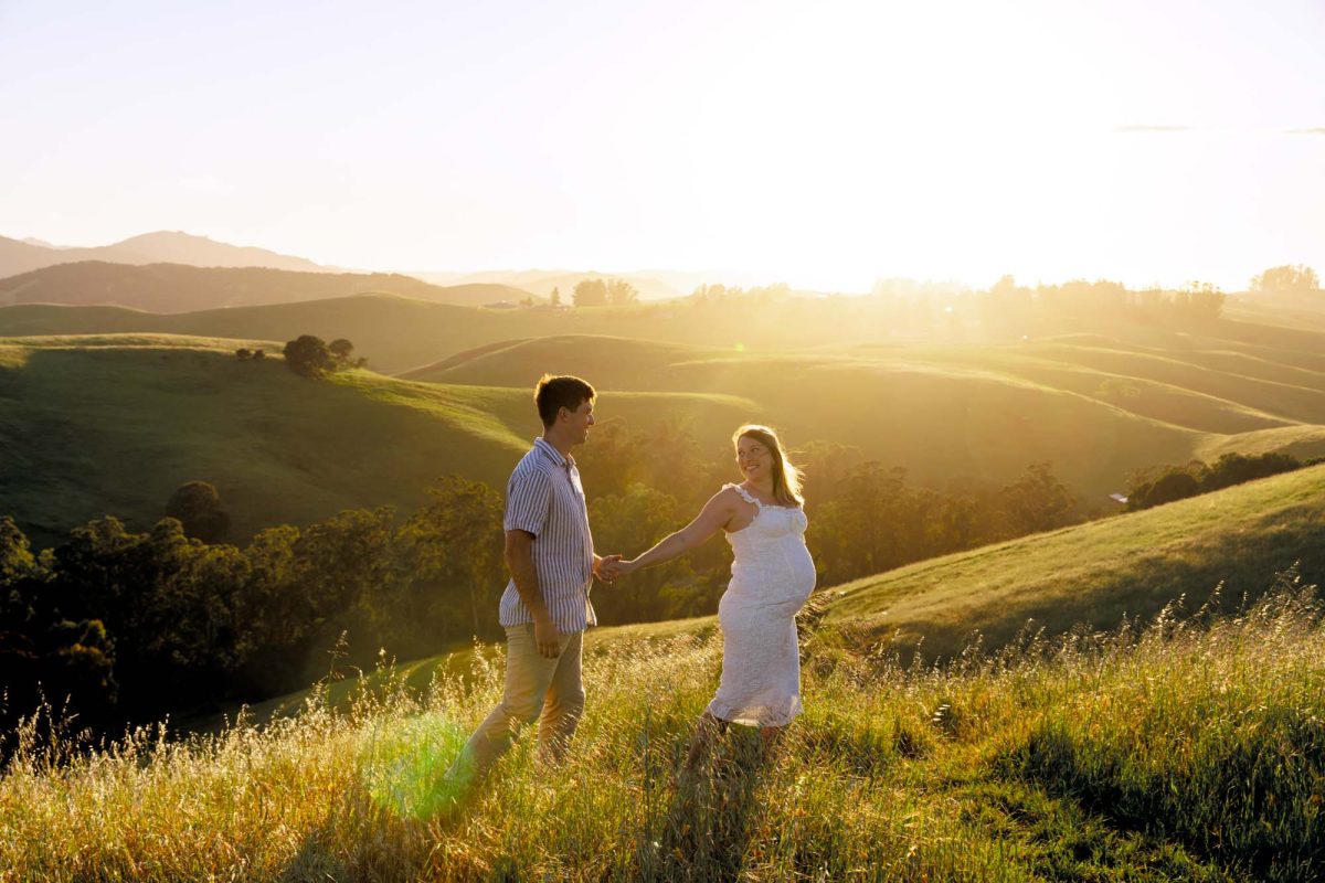 A pregnant woman in a white dress smiles with her hand on her belly as her partner holds her hand high during their Petaluma couples photo shoot. The couple stands in the tall grass of a sunlit hillside with golden sunset light illuminating the rolling hills behind them.