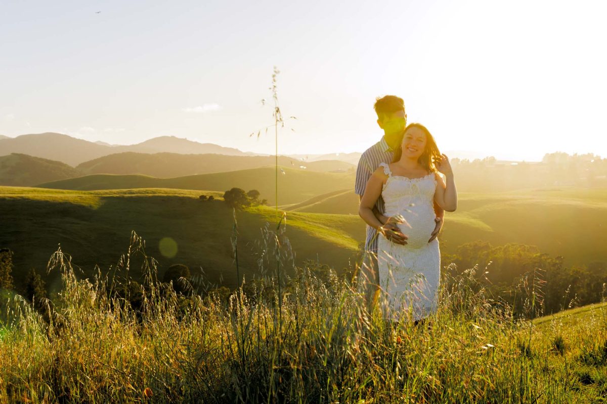 A smiling pregnant woman and her partner pose for their Petaluma maternity photography on a sunlit grassy hillside. He stands behind her with his arms around her and hands on her belly while she smiles toward the camera with one hand near her hair. Both are surrounded by tall grass as golden sunset light illuminates the rolling hills in the background. She wears a white dress and he wears a striped shirt.