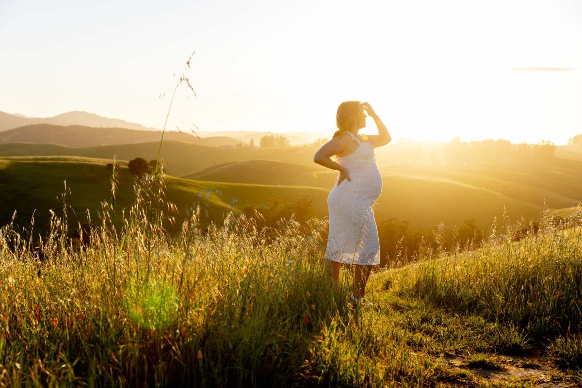 A pregnant woman poses for Petaluma maternity portraits on a sunlit grassy hillside. She stands in tall grass with one hand raised to fix her hair and the other resting on her hip while gazing out over the rolling hills. Warm golden sunset light bathes the scene and creates a bright glow around her. She wears a white dress.