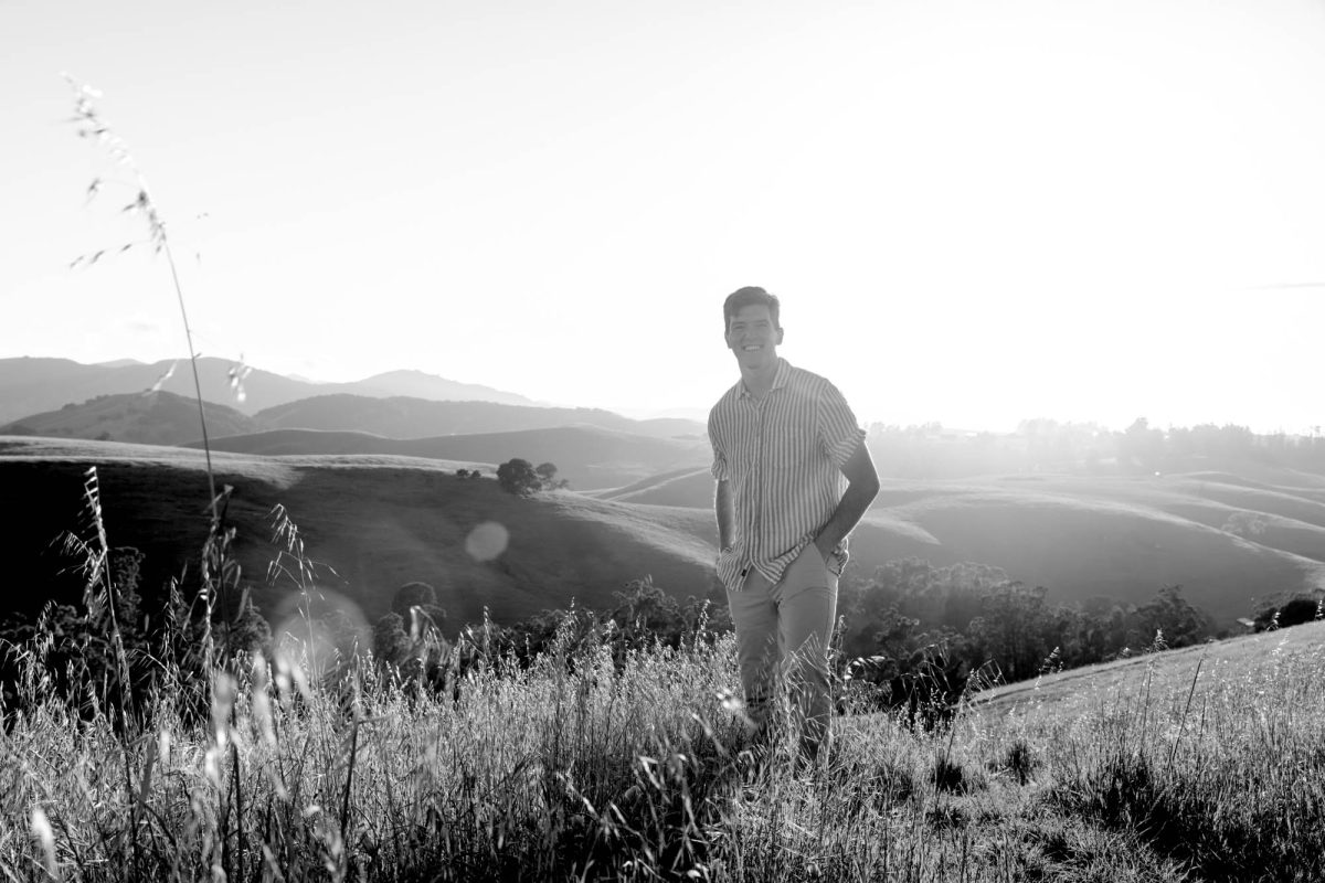 A smiling man in a striped shirt stands with his hands in his pockets in the tall grass as the Petaluma portrait photographer captures this man's portrait at Helen Putnam regional park. He faces the camera against the backdrop of rolling hills with bright backlighting in this black and white image.
