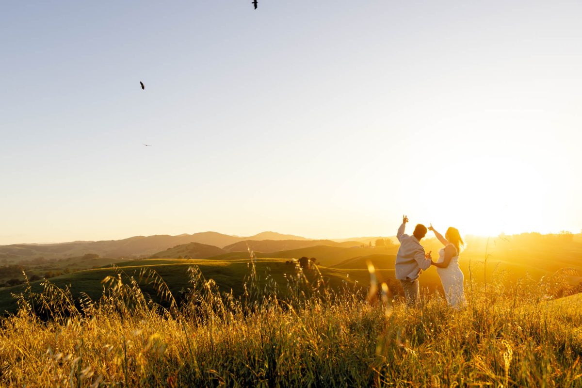 Petaluma portrait photographer Jason Guy captures a couple at the zenith of the sunset among grassy rolling hills. The man and woman both point up to the sky as birds fly through air above. The woman wears a white dress while the man wears a cream colored jacket and tan pants. 