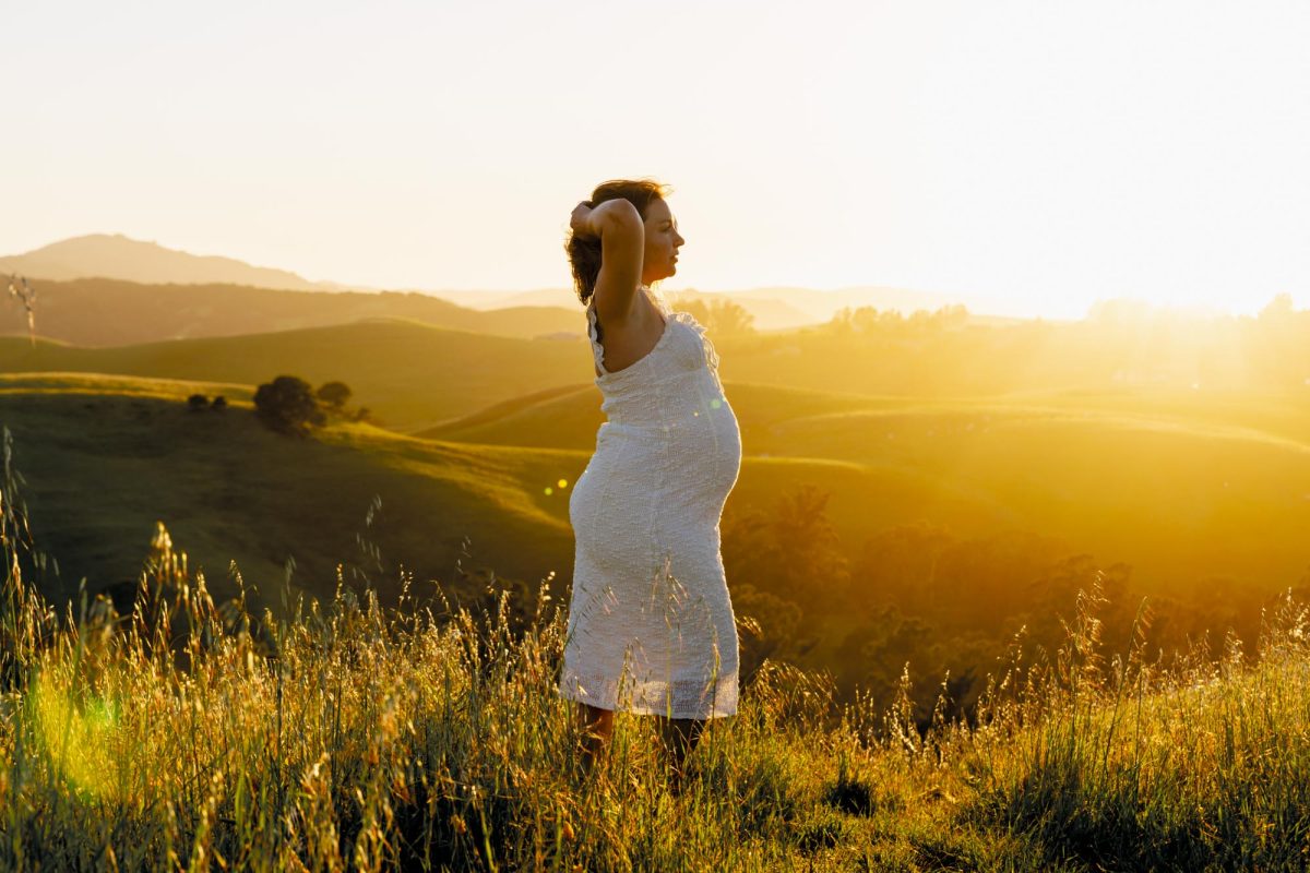 A pregnant woman poses for her Petaluma pregnancy photo shoot on a sunlit grassy hillside. She stands in tall grass with both hands behind her head while looking out over the rolling hills as warm golden sunset light bathes the scene. She wears a white dress.