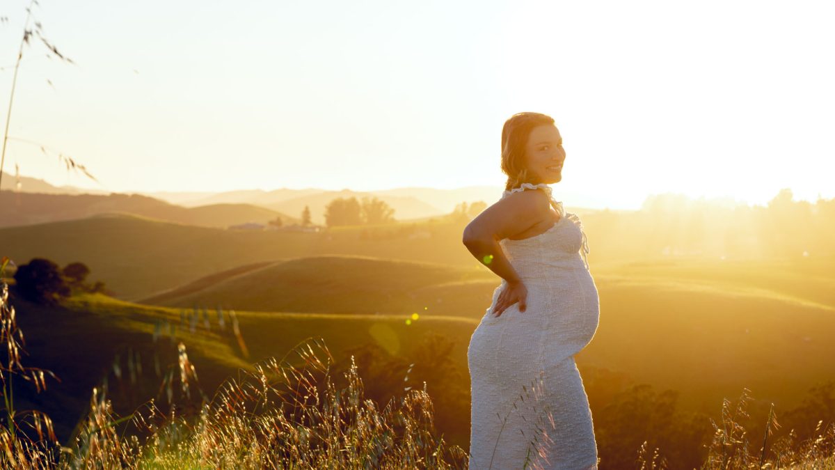 A pregnant woman poses for her Petaluma pregnancy photography session on a sunlit grassy hillside. She stands in tall grass with one hand on her hip and smiles over her shoulder toward the camera as warm golden sunset light bathes the rolling hills behind her. She wears a white dress.